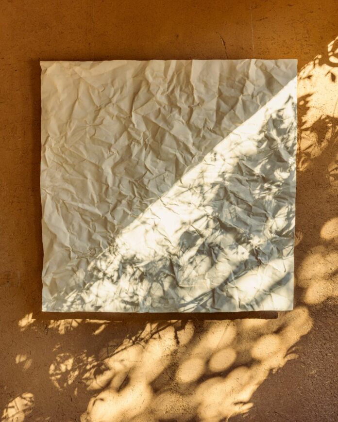 A square, wrinkled, off-white piece of paper is suspended on a textured adobe wall.