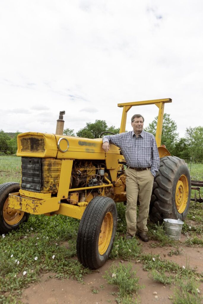 George Gundrey wearing a plaid shirt and tan pants is leaning against a vintage yellow tractor in a field.