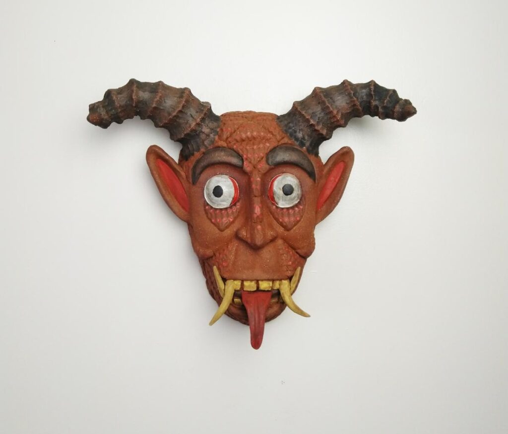 A brown ceramic mask of a devil's face with red-tipped ears, two white tusks, a red tongue sticking out, and two twisted, dark horns is shown against a white background.