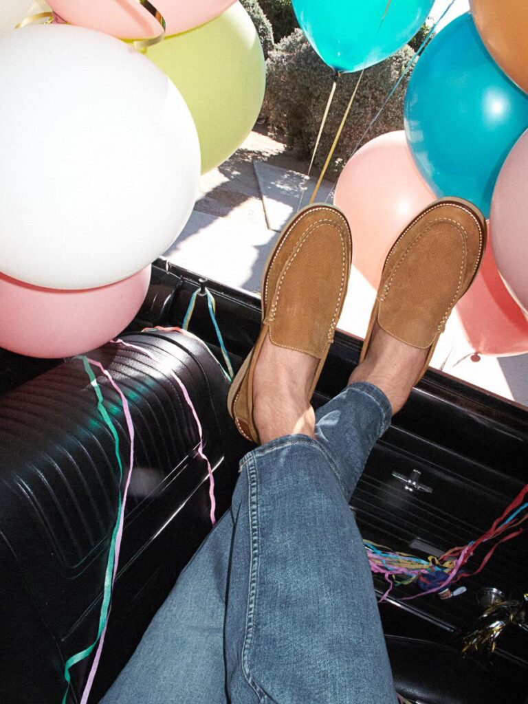 A man's feet wearing tan suede loafers are propped up on the dashboard of a car, surrounded by colorful balloons.
