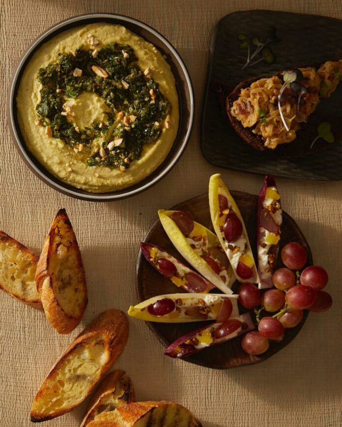 Three appetizers on a linen tablecloth: a bowl of white bean hummus topped with zhoug, endive leaves filled with grapes and cheese, and a piece of bread topped with a dip.