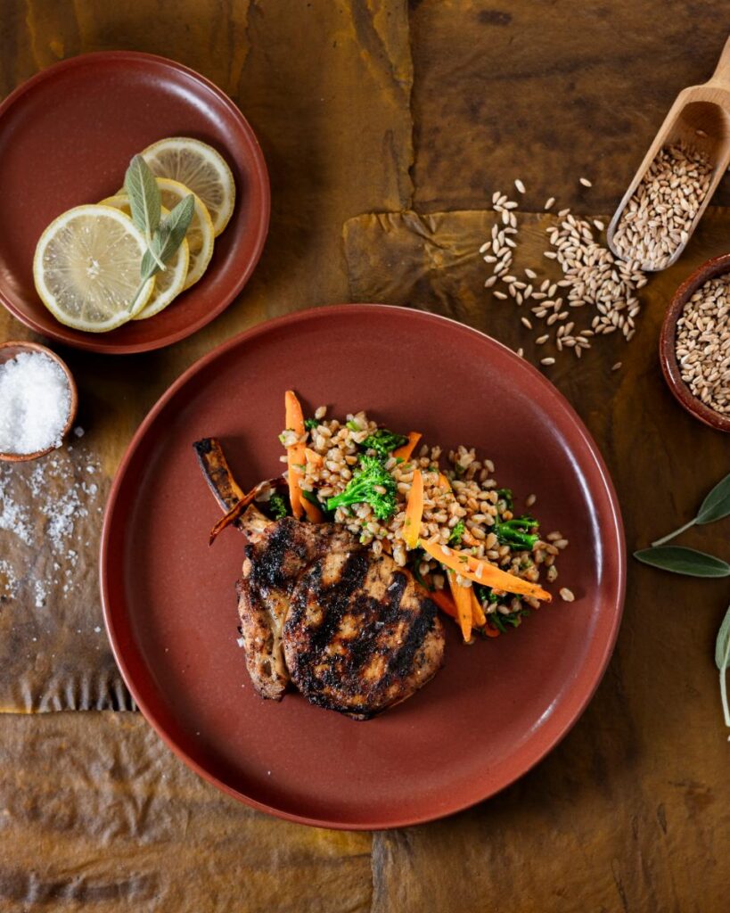 A grilled pork chop is on a red plate with farro, broccoli, and baby carrots, accompanied by a side plate of sliced lemons and sage leaves.