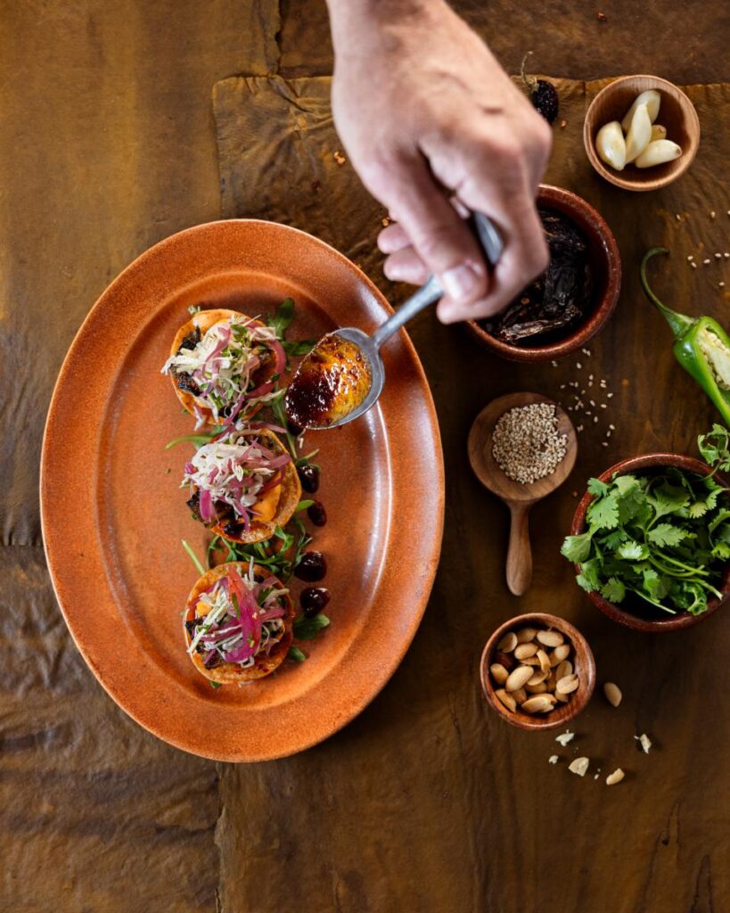 A hand holding a silver spoon drizzles salsa macha over three tostadas on a long, reddish-orange plate.