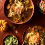 Two bowls of Haitian Pumpkin-Beef Soup are shown on a reddish-brown surface, garnished with shredded cabbage and green onions.