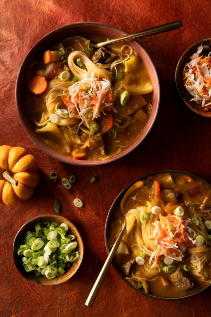 Two bowls of Haitian Pumpkin-Beef Soup are shown on a reddish-brown surface, garnished with shredded cabbage and green onions.