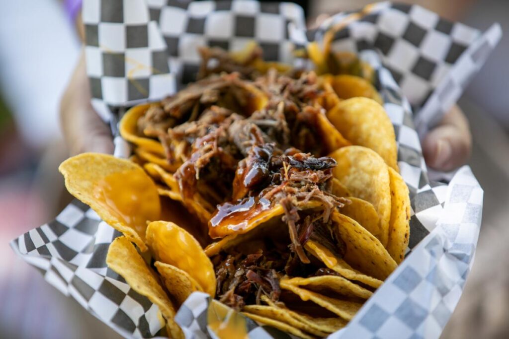A hand is holding a black and white checkered paper container filled with a pile of nachos topped with pulled pork and a drizzle of barbecue sauce.