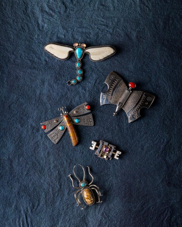 A flat-lay view of five pieces of silver jewelry shaped like insects, including a dragonfly with turquoise and bone wings and a spider with a turquoise stone, on a dark blue textured background.