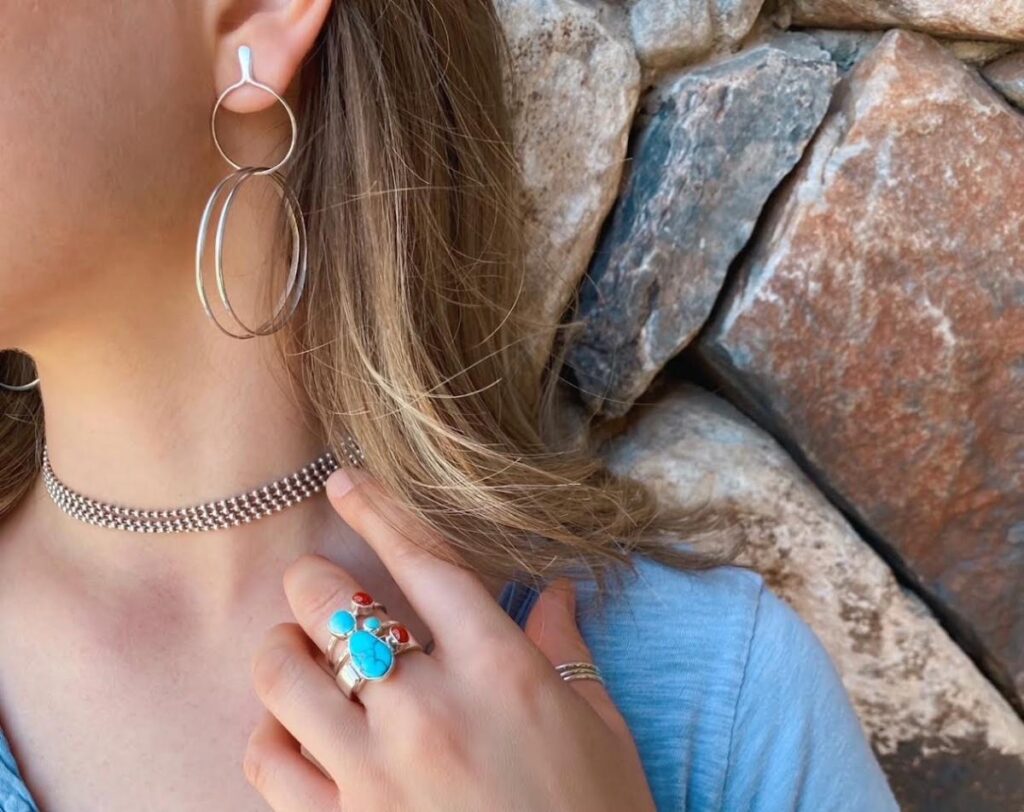 A pair of silver double hoop earrings are worn by a woman who is also wearing a silver choker and a silver ring set with turquoise and coral.