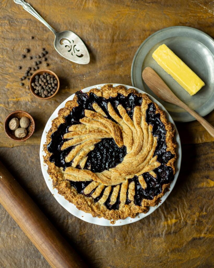 A dark purple huckleberry-merlot pie with a decorative woven crust is on a white plate.