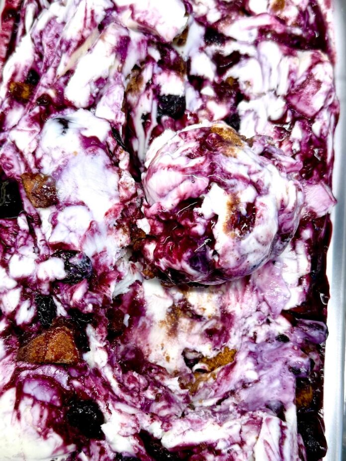 A close-up of a container of small-batch ice cream with swirls of purple berry and pieces of crumble, with a single scoop in the center.