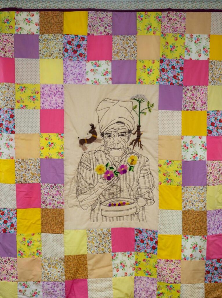 A quilt featuring a central embroidered image of an elderly man holding a bowl of flowers, surrounded by a border of multicolored, floral-patterned and solid fabric squares.
