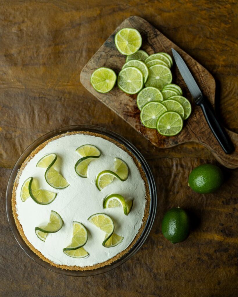 A whole key lime pie with a white topping and twists of lime zest is on a wooden surface next to a cutting board with a pile of sliced limes and a knife.