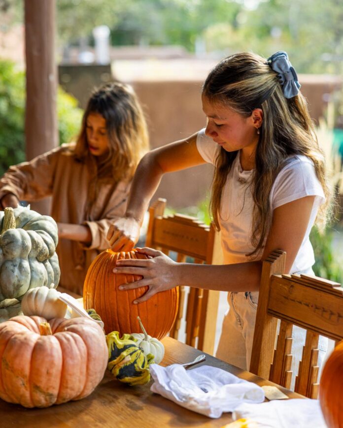 Two young girls are carving pumpkins on a wooden table outdoors, with a pile of various gourds and pumpkins next to them.