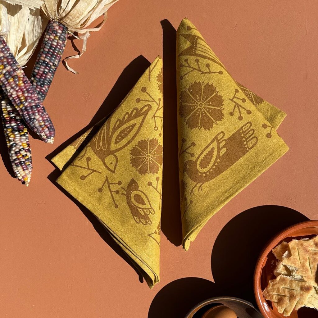 A table setting featuring two gold napkins with a bird and flower design, and a bowl of flatbread.