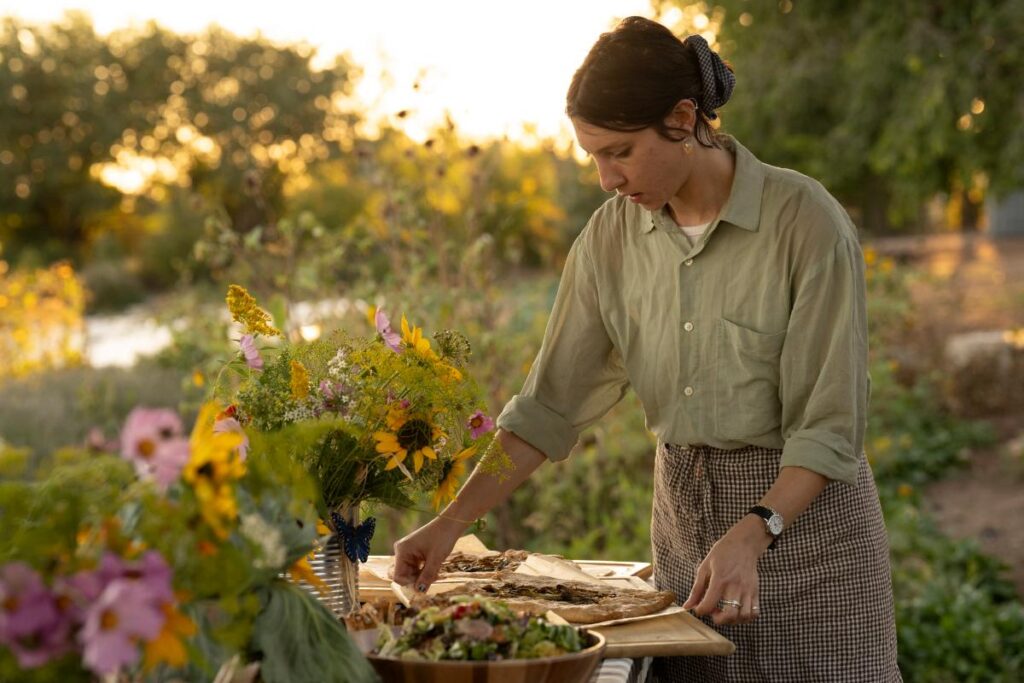 A woman with a bun and a polka dot hair tie, wearing a checkered apron, stands at a long wooden table, reaching for a slice of food from a tray of a seared leek galette.