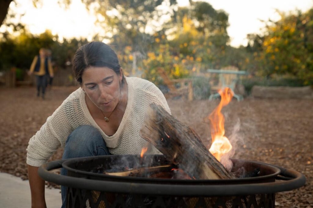 A woman with long dark hair, wearing a white sweater, crouches next to a metal fire pit, blowing on a burning log to encourage the flames.