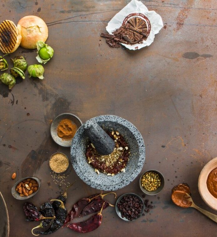 A flat lay photo showing ingredients for Mexican mole sauce, including various chiles, nuts, spices, and a stone mortar and pestle on a rustic metal surface.