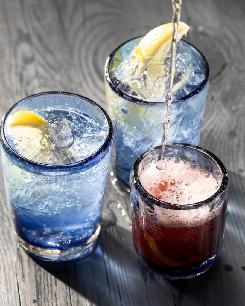 A pair of mouth-blown, blue-rimmed drinking glasses are shown, with a smaller blue-rimmed glass next to them as a drink is poured.
