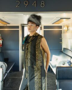 Native Fashion 32 Week 2025 TABLE Magazine A person in a pantssuit and fluffy hat poses between benches of chairs.