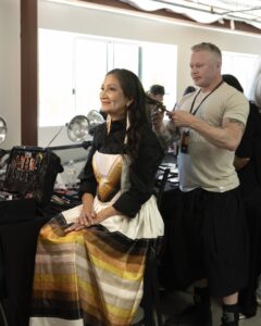 Native Fashion Week 2025 15 TABLE Magazine A woman with long dark hair wears an orange, yellow, brown, and white dress.