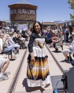 Native Fashion Week 2025 17 TABLE Magazine A woman in a white, yellow, orange, and brown dress poses on an outside runway at Santa Fe Railyard.