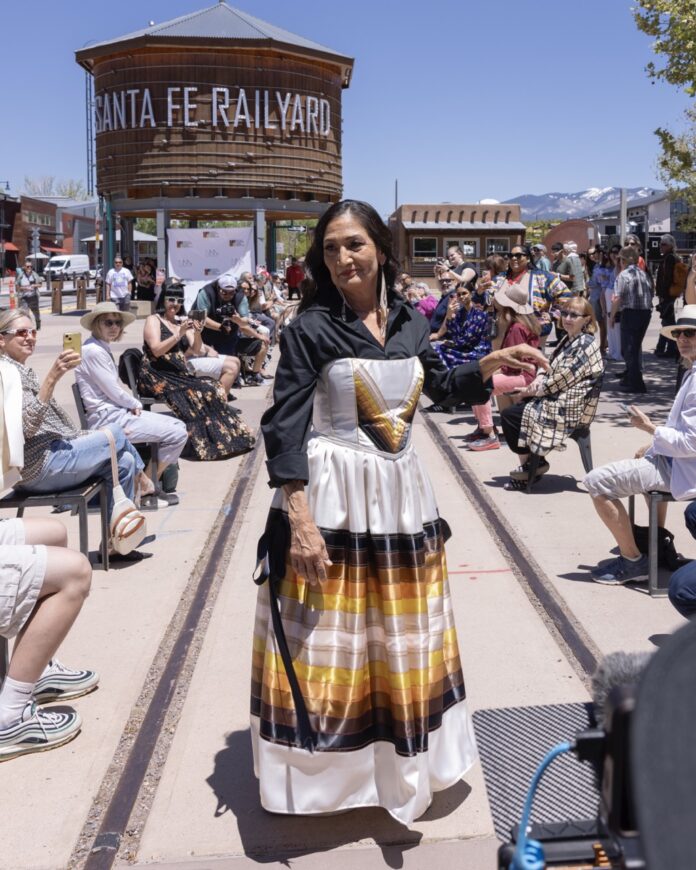 A woman in a white, yellow, orange, and brown dress poses on an outside runway at Santa Fe Railyard.