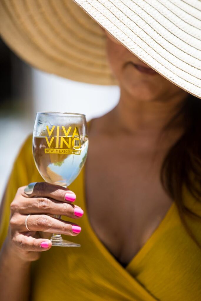 A woman wearing a wide-brimmed straw hat is holding a glass of white wine with "VIVA VINO NEW MEXICO WINE" printed on it.
