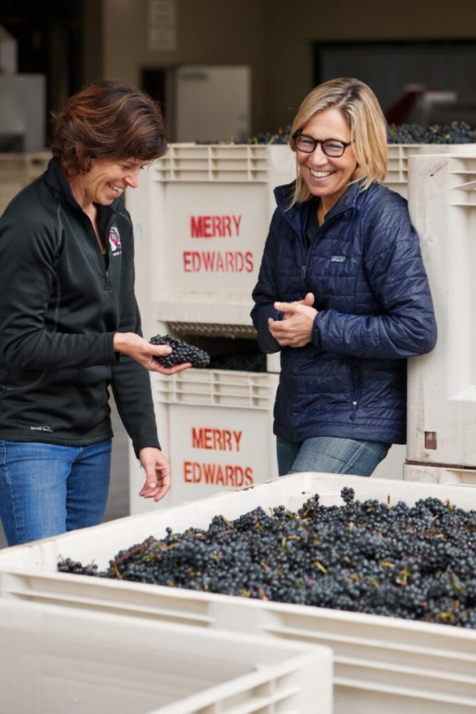 Two women, one holding a small bunch of dark grapes, are smiling while standing next to large bins of grapes at a winery.