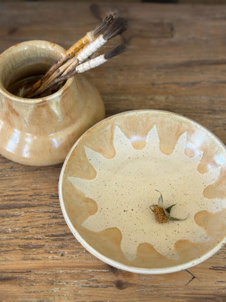 A ceramic plate with a textured, sunburst-like design in a beige color, next to a matching vase holding several paint brushes with wooden handles and deer bristles.