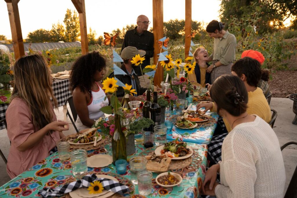 A large group of friends and family sitting around a long, outdoor table on a farm at sunset, sharing a feast.