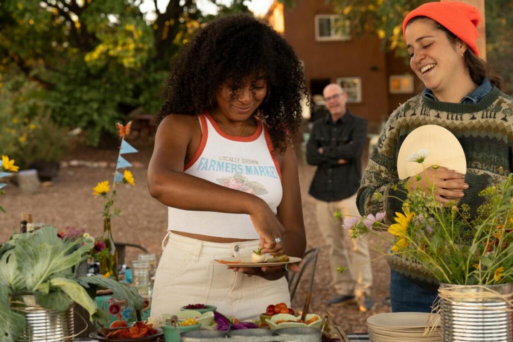 A smiling Black woman wearing a white tank stands at an outdoor table and dips a cracker into a bowl of dip. She is looking down at the food while a second woman in a sweater and orange beanie laughs, holding a wooden paddle with a white flower on it.
