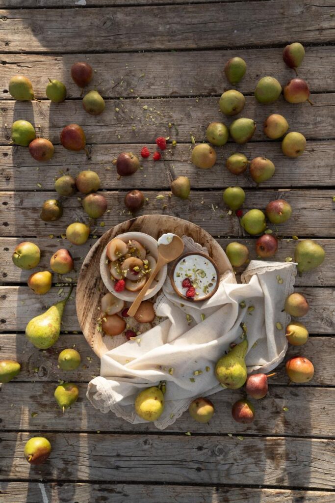 A rustic wooden table with a large round wooden platter holding a bowl of poached pear halves and a separate bowl of crème fraîche, both garnished with raspberries and chopped pistachios.
