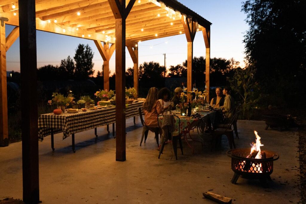 A group of people enjoying a dinner at a long table underneath a lit, rustic wooden pergola at sunset. A fire pit glows to the right of the table.
