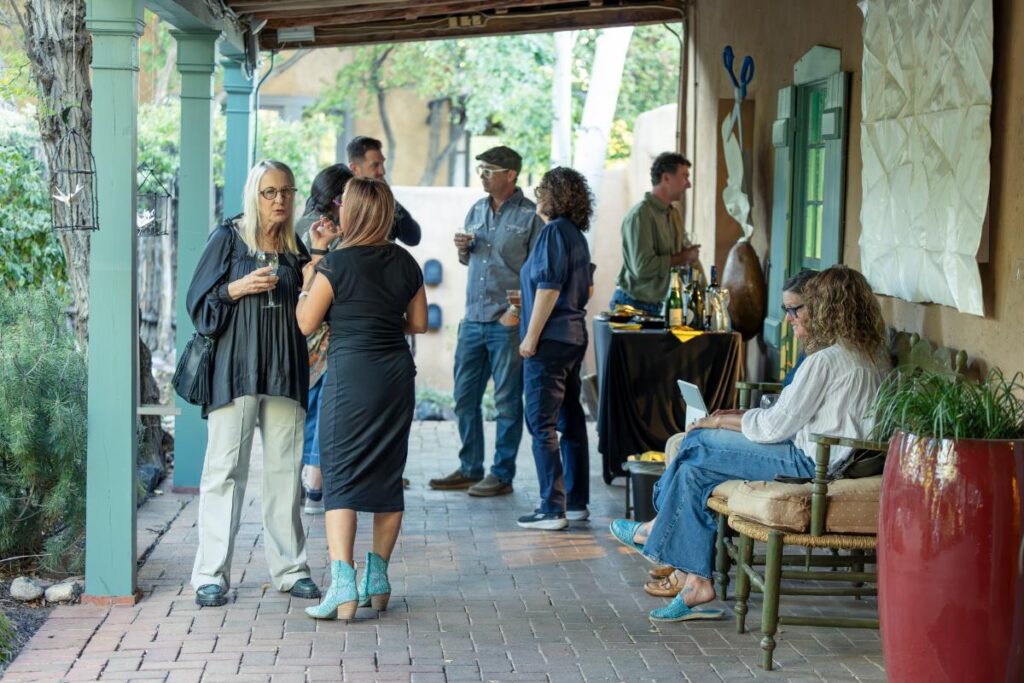 A large group of people mingling in a lush, outdoor space at sunset, holding drinks and chatting.