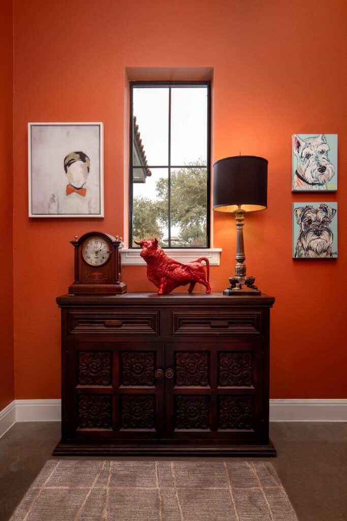 A wide shot of a mudroom with vibrant orange walls. A dark wooden cabinet with intricate carvings is placed below a small, rectangular window. A wooden clock sits on the left side of the cabinet, and a red bull statue stands in the center.
