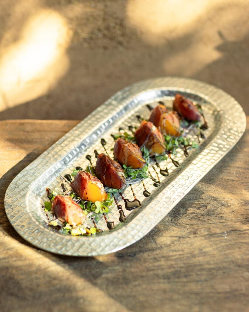 A close-up of a silver, hammered metal tray with six prosciutto-wrapped appetizers arranged on a bed of greens.