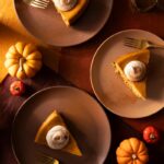 Three slices of pumpkin cheesecake on brown plates are arranged on a multicolored surface, each slice topped with a dollop of whipped cream and a sprinkle of cinnamon.