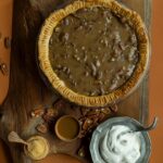 A baked pecan pie with a caramel topping is shown from above, set on a wooden cutting board.