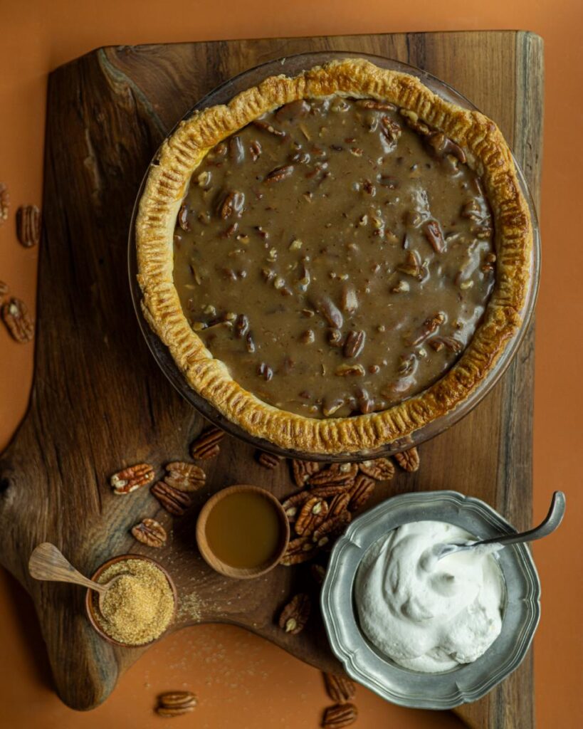 A baked pecan pie with a caramel topping is shown from above, set on a wooden cutting board.