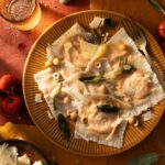 A plate of pumpkin-stuffed ravioli is garnished with brown butter, sage, hazelnuts, and shaved cheese.