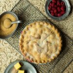 A whole baked raspberry rhubarb pie with a latticed, sugared crust is on a wire rack, with a pewter bowl of fresh raspberries and a small bowl of turbinado sugar nearby.