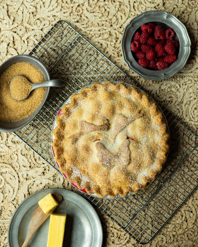 A whole baked raspberry rhubarb pie with a latticed, sugared crust is on a wire rack, with a pewter bowl of fresh raspberries and a small bowl of turbinado sugar nearby.
