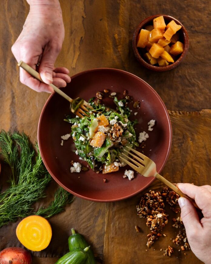 A person's hands holding gold-colored forks are serving a roasted beet salad with greens, granola, and tofu feta from a red bowl.
