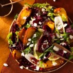 A colorful roasted pumpkin salad is presented on a plate with red and green lettuce, goat cheese, and pomegranate seeds.