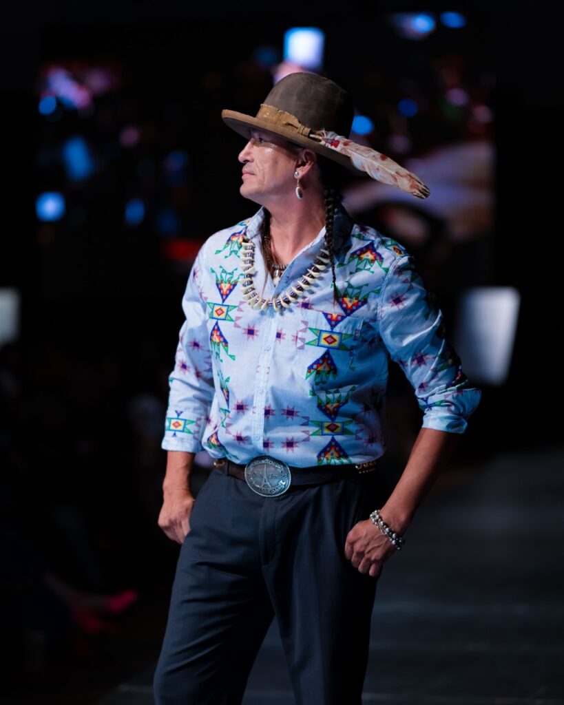 SWAIA Native Fashion Week 13 TABLE Magazine A man in a native shirt and hat with a feather poses on stage.