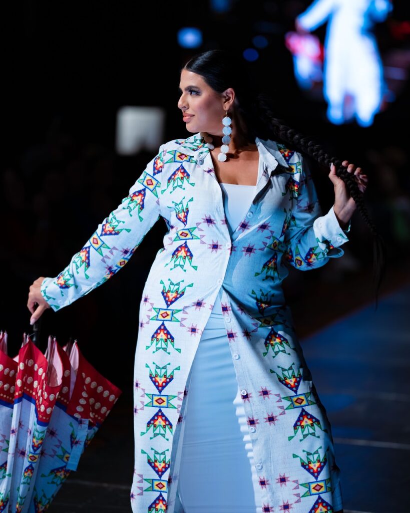 SWAIA Native Fashion Week 6 TABLE Magazine A woman in a blue dress and blue button up poses on stage.