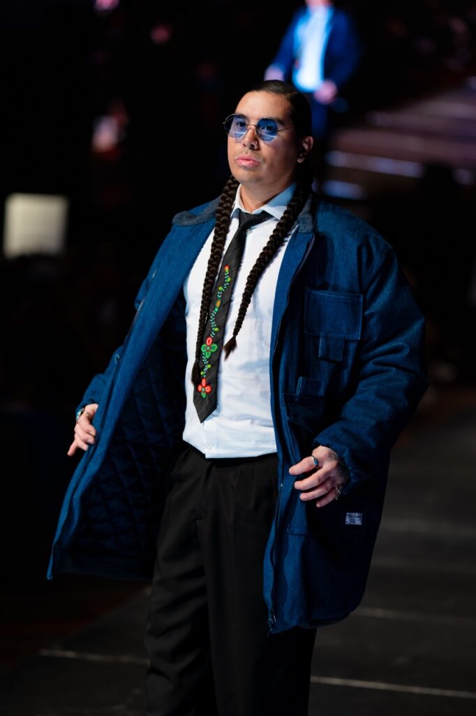 SWAIA Native Fashion Week 7 TABLE Magazine A man in a dark blue jacket and braids walks down a runway.