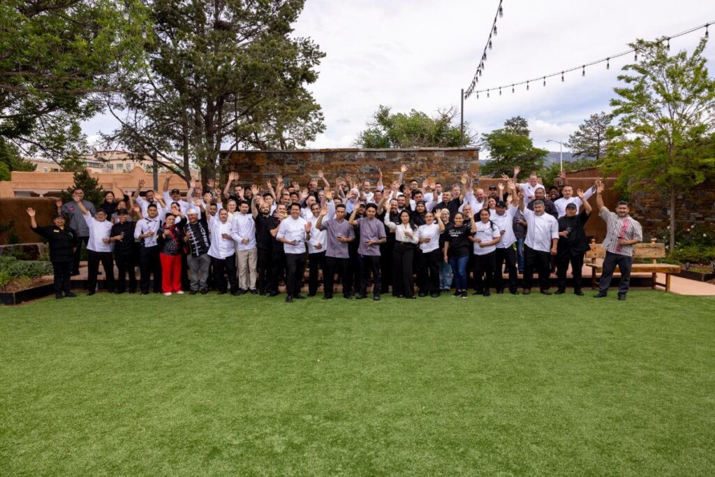 A large group of people, including chefs and event staff, are standing on a grassy lawn with their hands raised at Santa Fe Wine & Chile Fiesta.