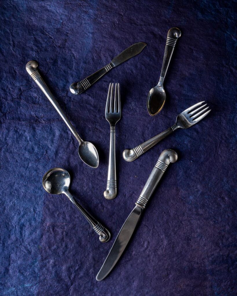 A high-angle view of a set of six antique silver forks, spoons, and knives with horizontal etched markings on the handles, arranged on a dark purple surface.