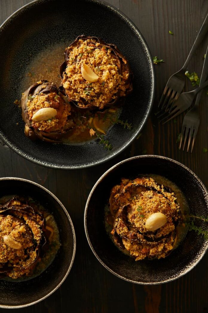 Three stuffed artichokes in black bowls, with a garlic clove garnish and a sauce in the bottom of the bowl.